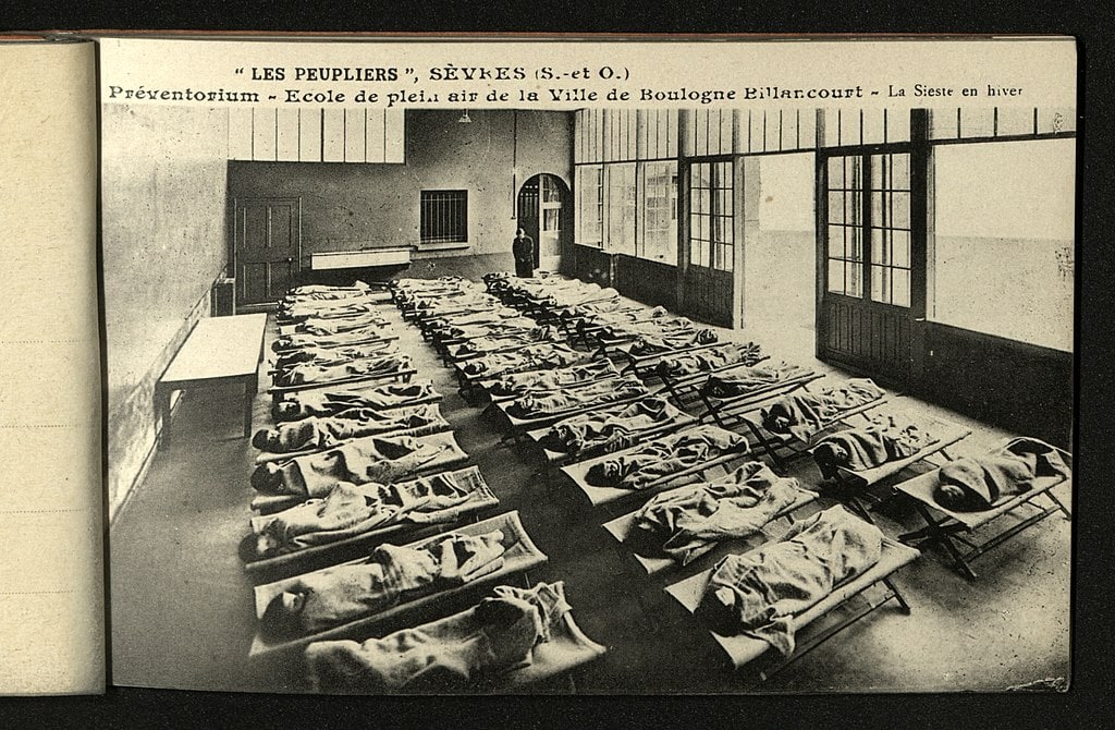 Two rows of children lying on cots in front of open windows at tuberculosis preventorium