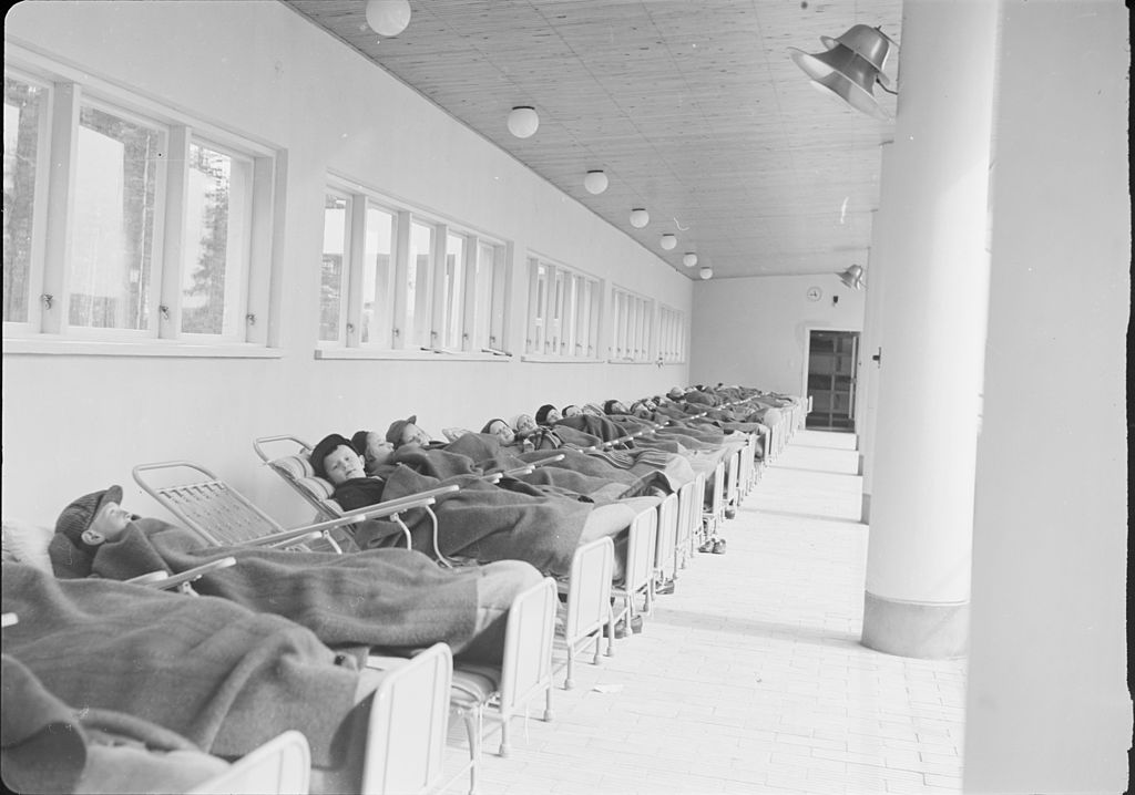 Children lying on beds lined up on an open-air porch.